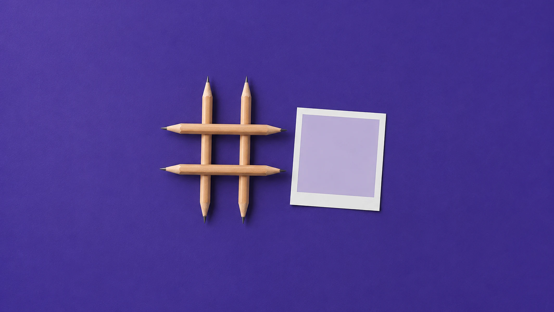 Quatro lápis de madeira cruzados formando o símbolo da hashtag, ao lado de uma foto quadrada em branco, sobre papel violeta