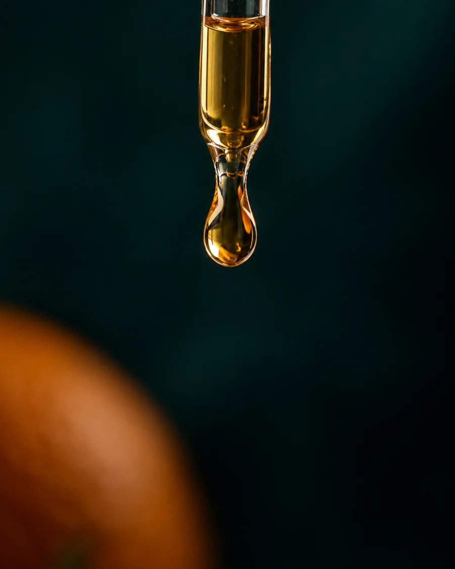 Uma gota dourada de sérum na ponta de um conta-gotas de vidro, sobre um fundo verde-escuro